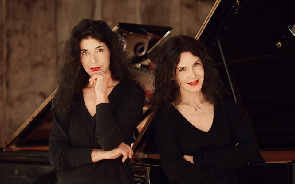 The pianists Katia and Marielle Labeque. The two women with long black hair and red lipstick look directly into the camera. In the background there are two Steinway Grand Pianos.