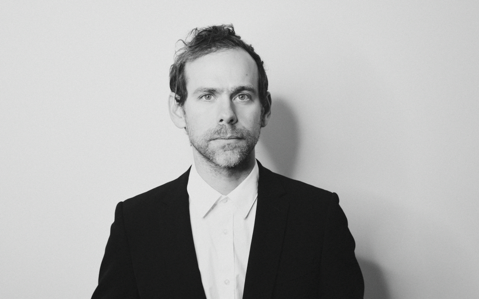 Photo of Bryce Dessner, credit Shervin Lainez. Image description: a black and whiteheadshot of the composer Bryce Dessner with a plain white background. A man with light skin wearing a shirt and suit jacket. He looks directly into the camera.